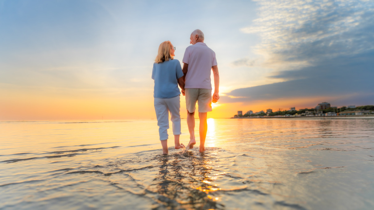 Achterkant van een ouder echtpaar dat hand in hand over het strand loopt tijdens hun vakantie bij zonsondergang.