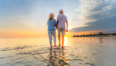 Achterkant van een ouder echtpaar dat hand in hand over het strand loopt tijdens hun vakantie bij zonsondergang.