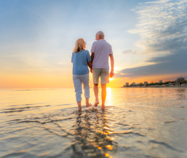 Achterkant van een ouder echtpaar dat hand in hand over het strand loopt tijdens hun vakantie bij zonsondergang.