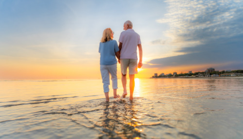 Achterkant van een ouder echtpaar dat hand in hand over het strand loopt tijdens hun vakantie bij zonsondergang.