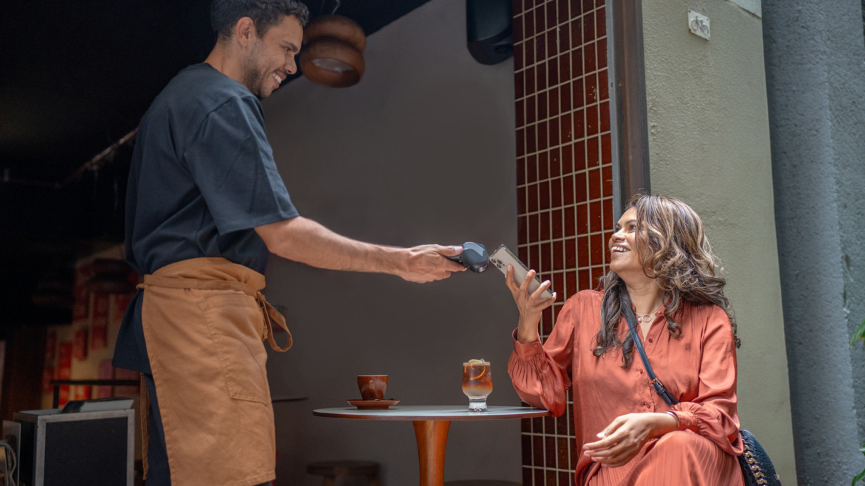 Mannelijke ober in Australië geeft glimlachend pinapparaat aan vrouwelijke klant op het terras