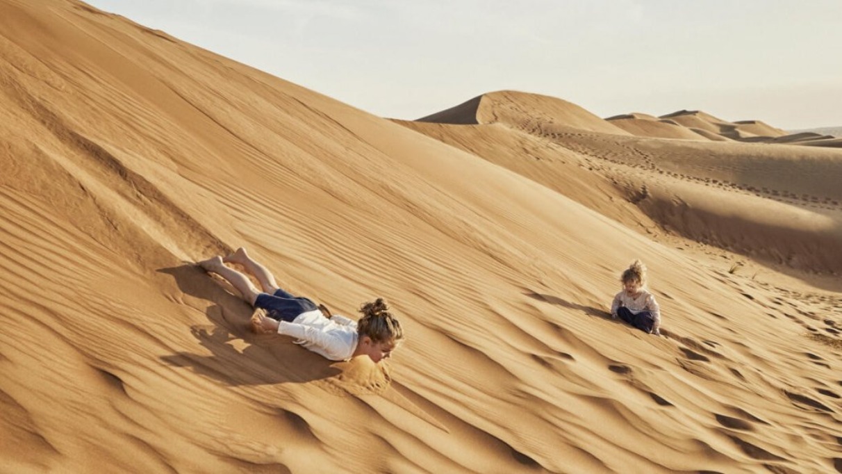 Twee kinderen spelen en glijden van een zandduin in een uitgestrekte woestijn, omringd door glooiende zandheuvels.