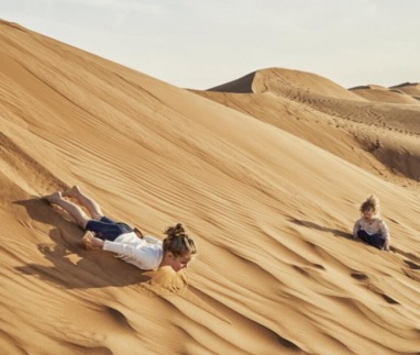 Twee kinderen spelen en glijden van een zandduin in een uitgestrekte woestijn, omringd door glooiende zandheuvels.