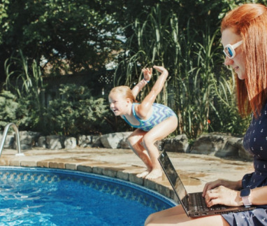Vrouw met laptop werkt naast een zwembad, terwijl een kind in het water springt, omringd door groen en bloemen.