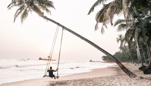 Reiziger schommelt aan een palmboom op een tropisch strand in Ghana, met de oceaan op de achtergrond.