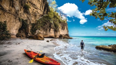 Een reiziger wandelt langs een afgelegen strand op Dominica, met een kajak op het zand en indrukwekkende kliffen op de achtergrond.