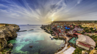 Adembenemend uitzicht op het kleurrijke kustdorp Popeye Village en Anchor Bay en helder turquoise water in Malta, omringd door dramatische rotsen en een gouden zonsondergang.