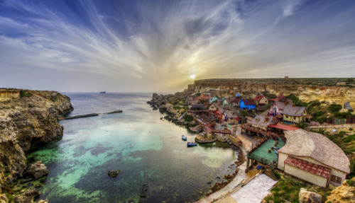 Adembenemend uitzicht op het kleurrijke kustdorp Popeye Village en Anchor Bay en helder turquoise water in Malta, omringd door dramatische rotsen en een gouden zonsondergang.