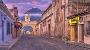 De iconische gele boog van Santa Catalina in Antigua, Guatemala, Unesco-werelderfgoed, met de imposante Agua-vulkaan op de achtergrond bij zonsondergang.