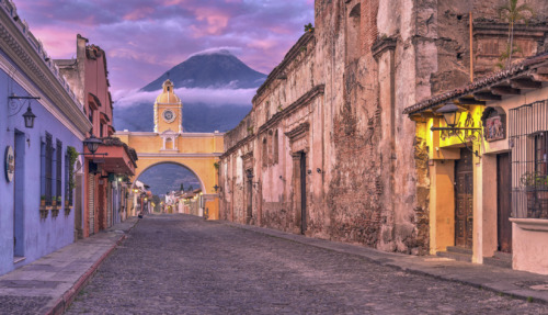 De iconische gele boog van Santa Catalina in Antigua, Guatemala, Unesco-werelderfgoed, met de imposante Agua-vulkaan op de achtergrond bij zonsondergang.