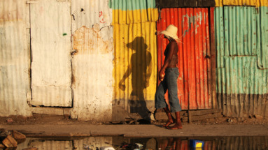 Een vrouw met een strohoed loopt langs een muur van kleurrijke golfplaten in Haïti, weerspiegeld in een plas water.