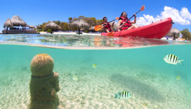 Twee mensen kajakken op het heldere water van Honduras, met een onderwaterbeeld van vissen en Mayaruïnes op de zeebodem.