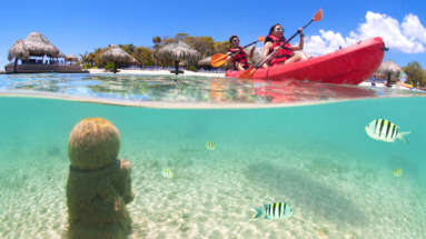Twee mensen kajakken op het heldere water van Honduras, met een onderwaterbeeld van vissen en Mayaruïnes op de zeebodem.