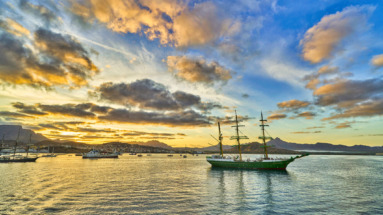 Historisch zeilschip in de haven van Kaapverdië bij zonsondergang, met bergen en een kleurrijke lucht op de achtergrond.