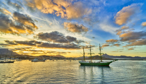 Historisch zeilschip in de haven van Kaapverdië bij zonsondergang, met bergen en een kleurrijke lucht op de achtergrond.