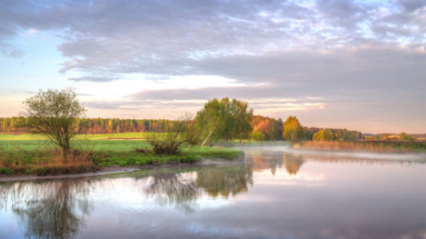 Een serene zonsopgang aan een rivier in Wit-Rusland, omgeven door groene velden en kleurrijke bomen.