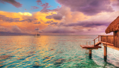 Overwaterbungalow met uitzicht op de turquoise lagune en een zeilboot bij zonsondergang in Frans-Polynesië