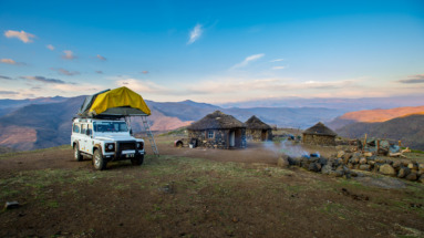 en kampeerauto met daktent naast traditionele rondavels in de bergen van Lesotho.