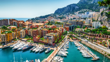 Haven van Monte Carlo vol luxe jachten omringd door kleurrijke gebouwen en bergen in Monaco, onder een heldere blauwe hemel.