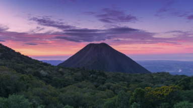 Uitzicht op de Izalco-vulkaan bij zonsondergang in El Salvador, omringd door groene heuvels en een kleurrijke hemel.