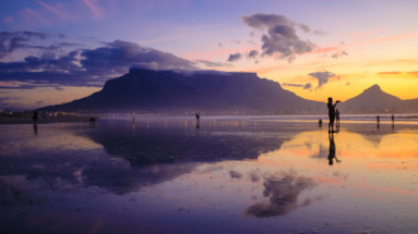 Een schilderachtige zonsondergang bij de Tafelberg in Kaapstad, Zuid-Afrika, met reflecties op het strand en silhouetten van mensen.