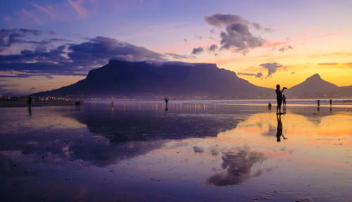 Een schilderachtige zonsondergang bij de Tafelberg in Kaapstad, Zuid-Afrika, met reflecties op het strand en silhouetten van mensen.