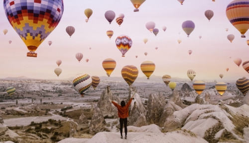 Kleurrijke luchtballonnen stijgen op boven de rotsformaties van Cappadocië bij zonsopgang, terwijl een reiziger het uitzicht bewondert.
