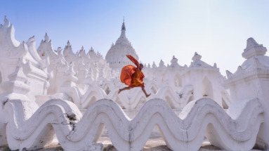 Een jonge monnik in oranje gewaad springt over de golvende witte structuren van de Hsinbyume-pagode in Myanmar.