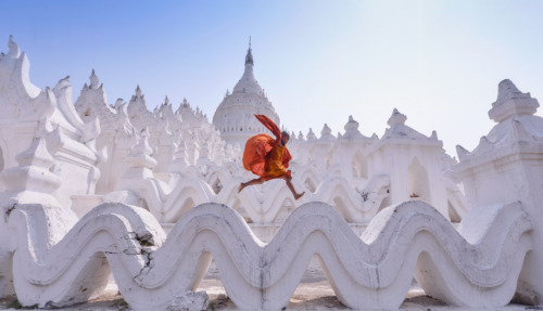 Een jonge monnik in oranje gewaad springt over de golvende witte structuren van de Hsinbyume-pagode in Myanmar.