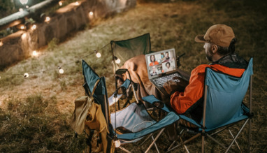 Man op campingstoel kijkt naar een online webinar op zijn laptop, omringd door sfeerverlichting.