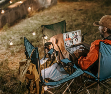 Man op campingstoel kijkt naar een online webinar op zijn laptop, omringd door sfeerverlichting.