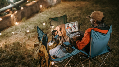 Man op campingstoel kijkt naar een online webinar op zijn laptop, omringd door sfeerverlichting.