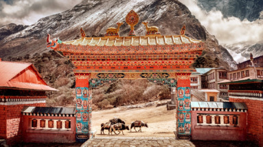 Een kleurrijke toegangspoort tot het Tibetaanse klooster met de besneeuwde Himalaya op de achtergrond en lastdieren op de voorgrond.