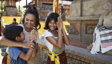 Een vrouw en kinderen in traditionele kleding begroeten elkaar met een glimlach bij een tempel.
