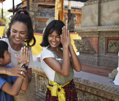 Een vrouw en kinderen in traditionele kleding begroeten elkaar met een glimlach bij een tempel.