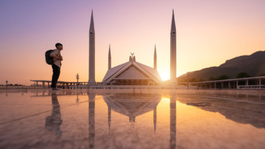De Faisal-moskee in Islamabad, Pakistan, bij zonsondergang met een reiziger op de voorgrond en reflectie in de marmeren vloer.