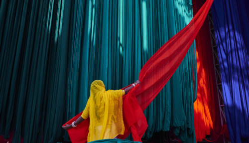 Een Indiase vrouw in een felgele sari hangt rood textiel op tegen een achtergrond van kleurrijke stoffen.