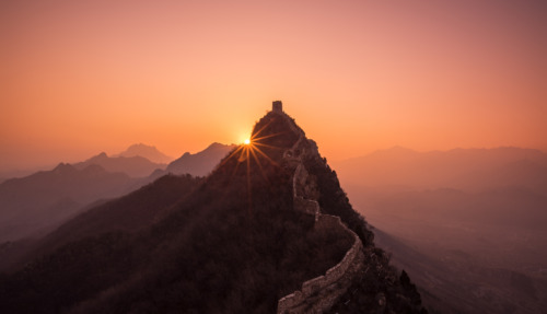 De Chinese Muur kronkelt over de heuvels, terwijl de zon opkomt achter een uitkijktoren en een magische gloed over het landschap werpt.