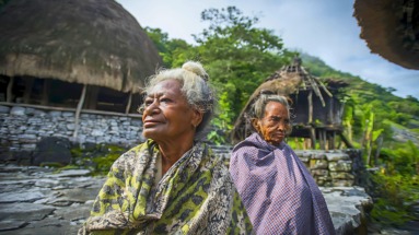 Twee oudere vrouwen in traditionele kleding in een dorp in Oost-Timor, omringd door traditionele hutten en groene natuur.
