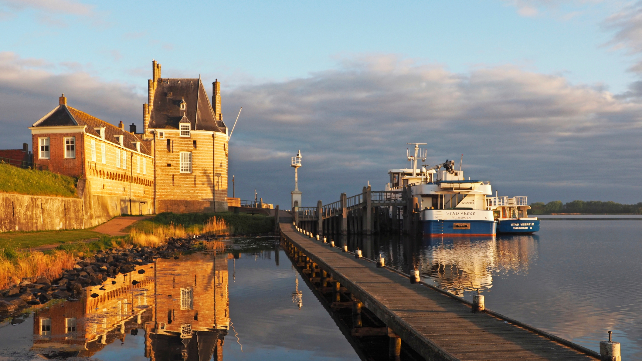 De historische Campveerse Toren in Veere, weerspiegeld in het water, met een steiger en veerboten in het avondlicht.