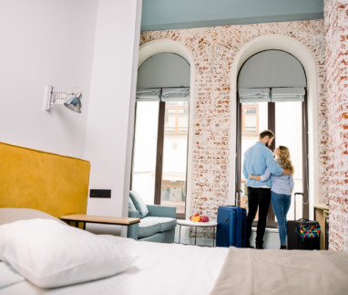 Een jong, vrolijk stel dat elkaar omhelst terwijl ze met grote koffers een hotelkamer binnenkomen, gezien van achteren.