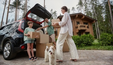 Gezin dat verhuisdozen uitlaadt bij hun nieuwe huis in een bosrijke omgeving, met een hond op de voorgrond.