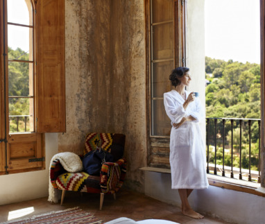 Vrouw in badjas geniet van een kopje koffie in een sfeervolle hoekkamer met grote ramen en een prachtig uitzicht op groene heuvels.