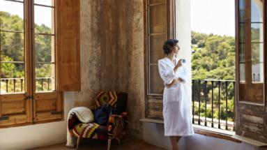 Vrouw in badjas geniet van een kopje koffie in een sfeervolle hoekkamer met grote ramen en een prachtig uitzicht op groene heuvels.