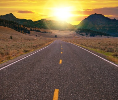 Straight two lane highway with no traffic leads through a valley landscape toward a golden sunrise over the horizon ahead. Conceptual for freedom, enjoying the journey, and a fresh new day.