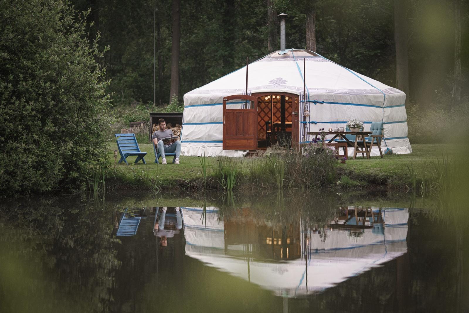 Traditionele witte yurt met houten deur aan de oever van een vijver in het Groningse bosgebied bij de Noorder Yurts, met een man zittend op een blauwe stoel.