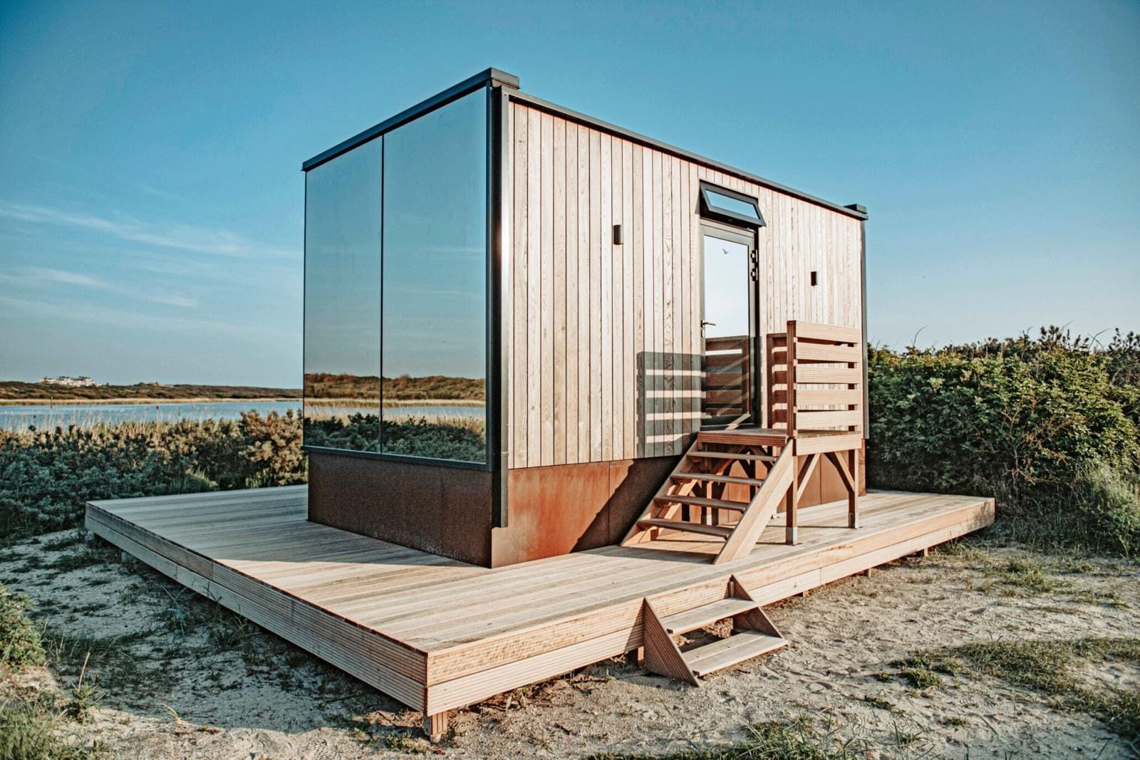 Moderne tiny house Base Camp met houten gevel en spiegelwand op een duinlocatie bij het water in IJmuiden, onder een blauwe hemel.