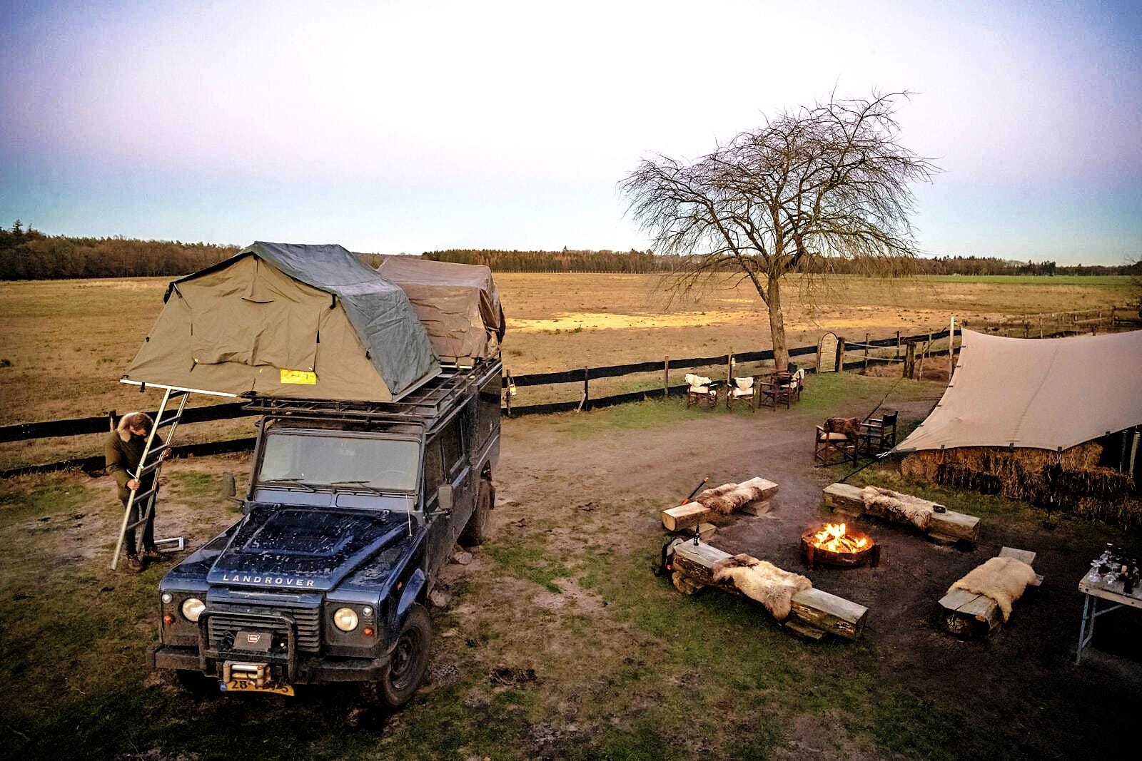Land Rover met daktent op een kampvuurplek in de natuur, met uitzicht op de Veluwse heidevelden bij zonsondergang.