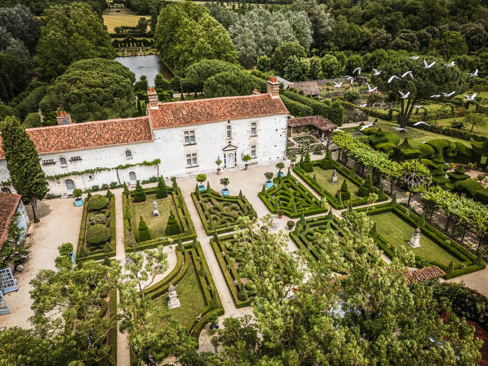 Luchtfoto van de symmetrische Franse tuinen van Les Jardins de William Christie in de Vendée, met kunstig gesnoeide hagen, beeldhouwwerken en een historisch landhuis