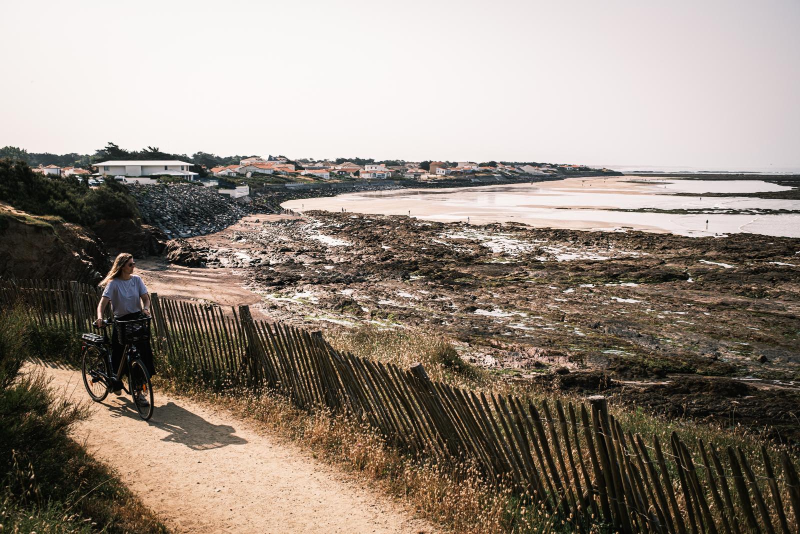 Vrouw fietst over een zandpad langs de rotsachtige kust en het strand van Brétignolles-sur-Mer in de Franse Vendée, met uitzicht op de zee en witte huisjes met rode daken.